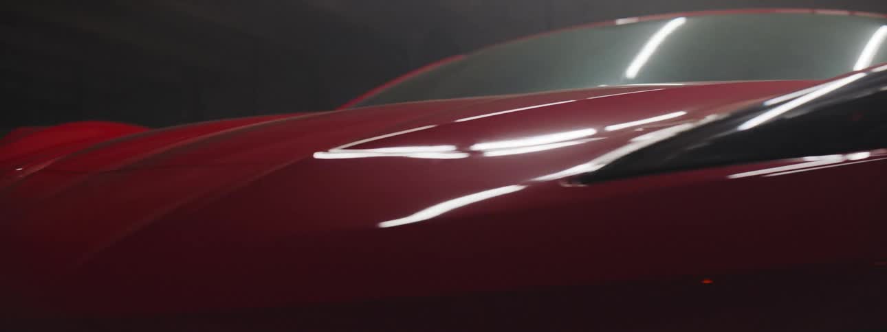 The front of a red sports car spins by on a floor display turntable in a dark room reflecting a distant florescent light