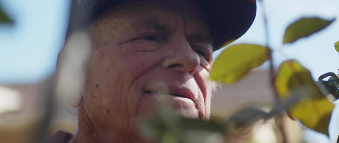 An elderly man is standing outdoors surrounded by nature