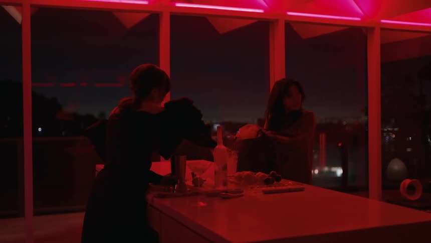 A woman is standing at a bar mixing a drink in a room with large windows and red light as another woman walks in with a large shoulder bag and puts on a poncho from the bag