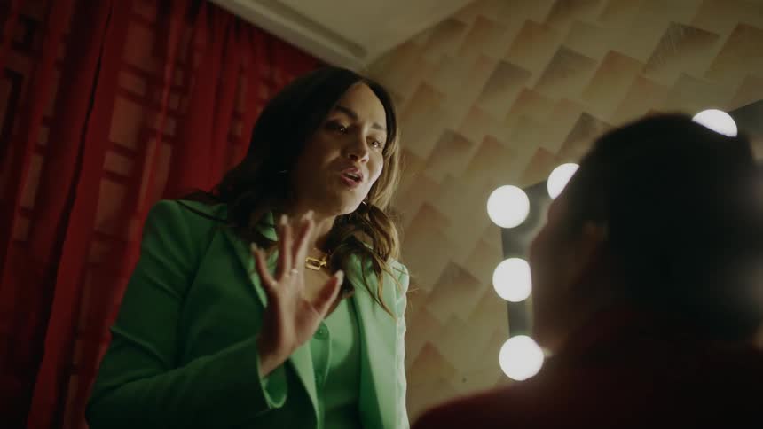 A woman in a green suit knelt down talking to another woman and then standing up while talking to her