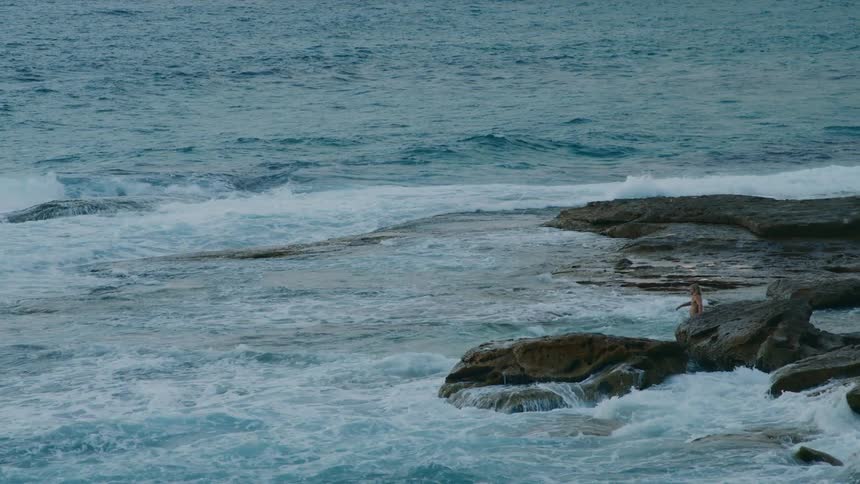 A man is walking across the beach near rocks as waves splash and roll in to shore