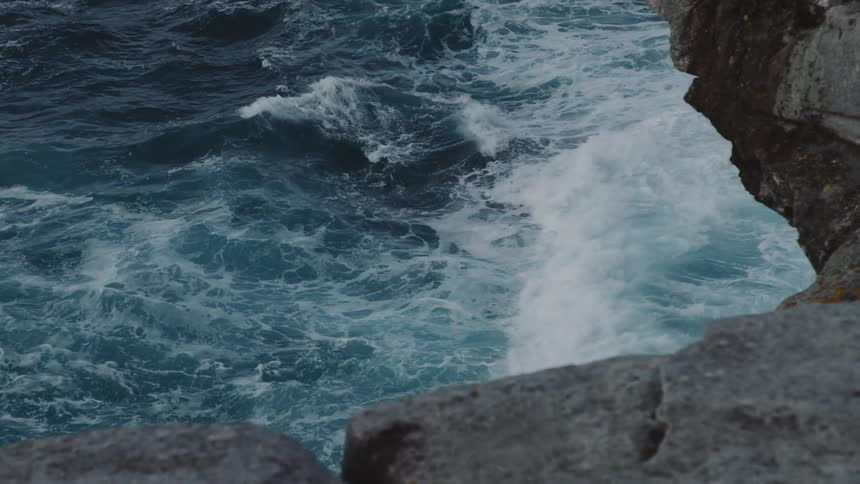 Waves crashing up against a coastline of rocks as foam forms on the surface of the water