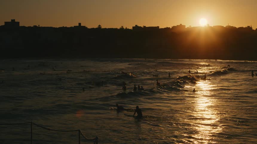 A large crowd of people swimming in sea against the coastal city at sunset