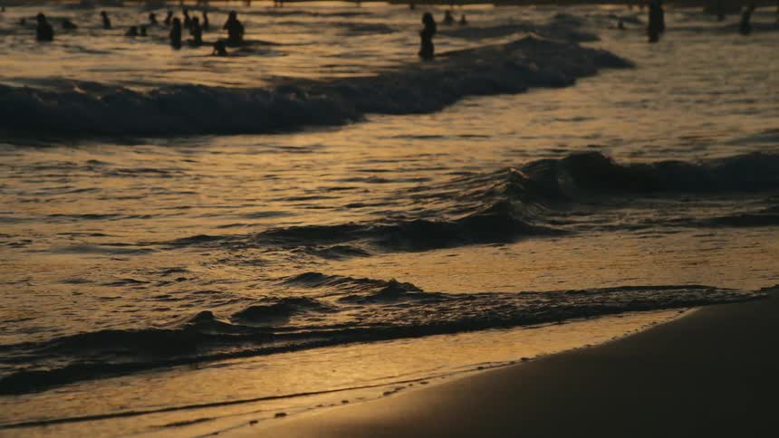 Waves crash against a beach at sunset while people swim in the background