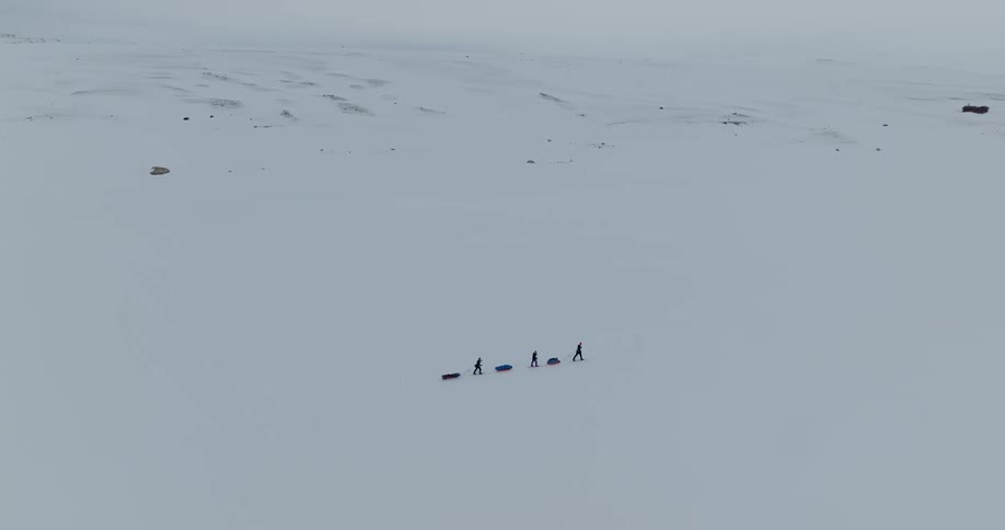 An aerial of three hikers walking through snow covered lands dragging sleds with their bags on them