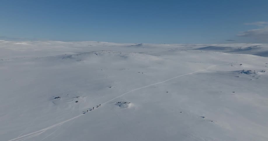 An aerial of wide open snowy landscapes as three people walk across the snow dragging sleds behind them