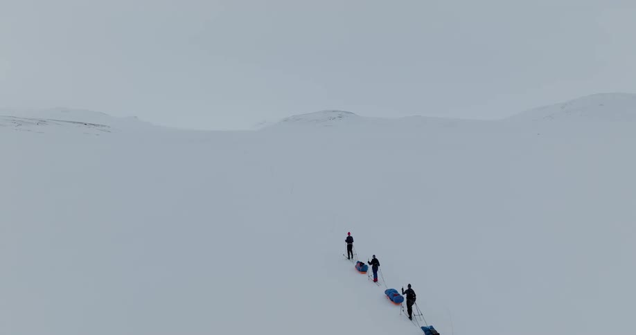 An aerial of three hikers walking through snow covered land dragging bags behind them