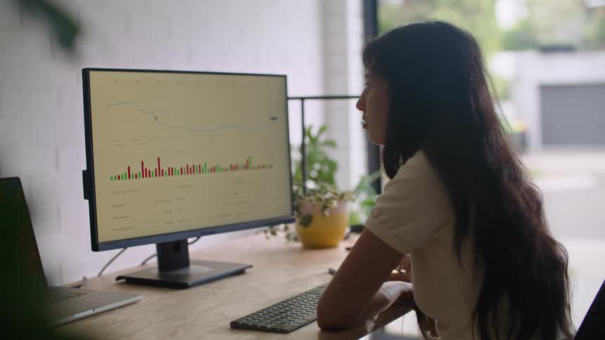 A teenage girl with long dark hair is sitting at a desk looking at a computer monitor with a moving graph on the screen