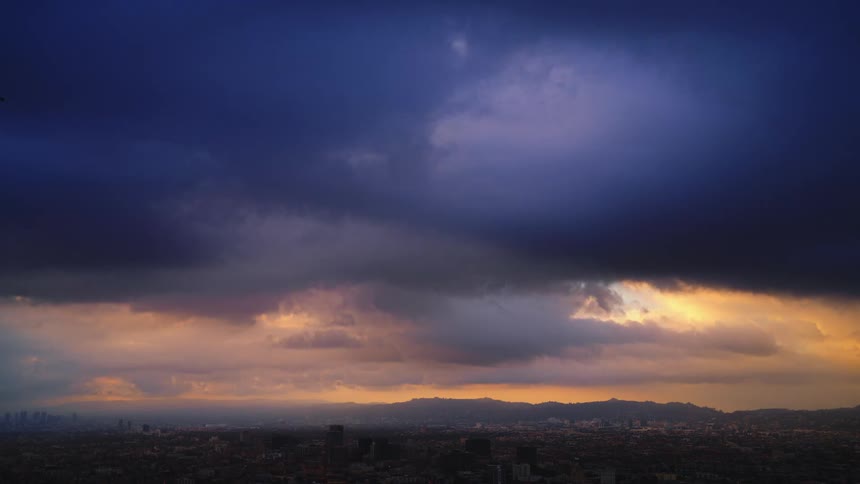 Clouds move over town and grow dark as the sun is setting in a colorful sky with mountains on the horizon