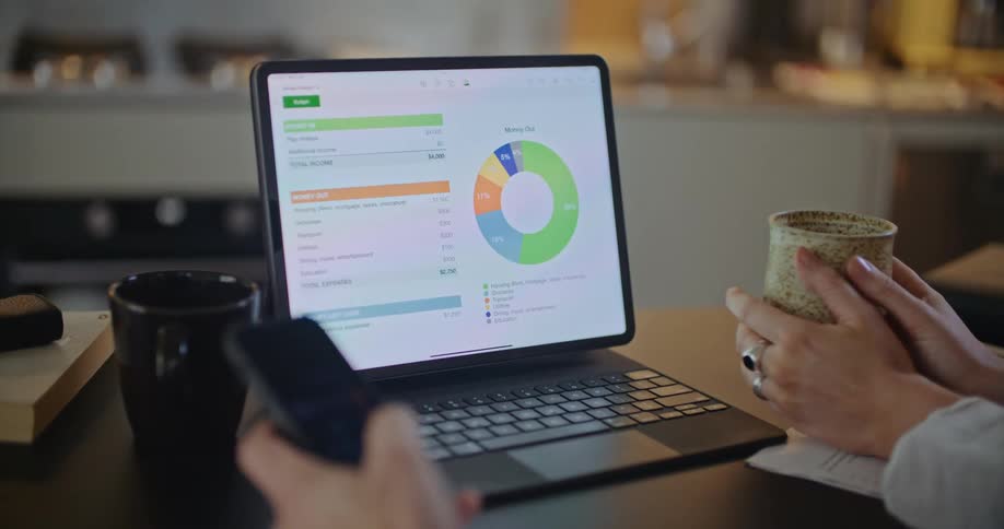 Man and woman sitting together at table looking at donut chart and finances on laptop while using smart phone and drinking coffee