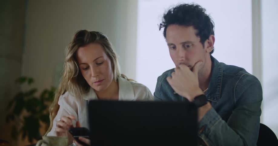 A man and woman sit next to each other while the girl looks at her cell phone and the man looks and points at the laptop