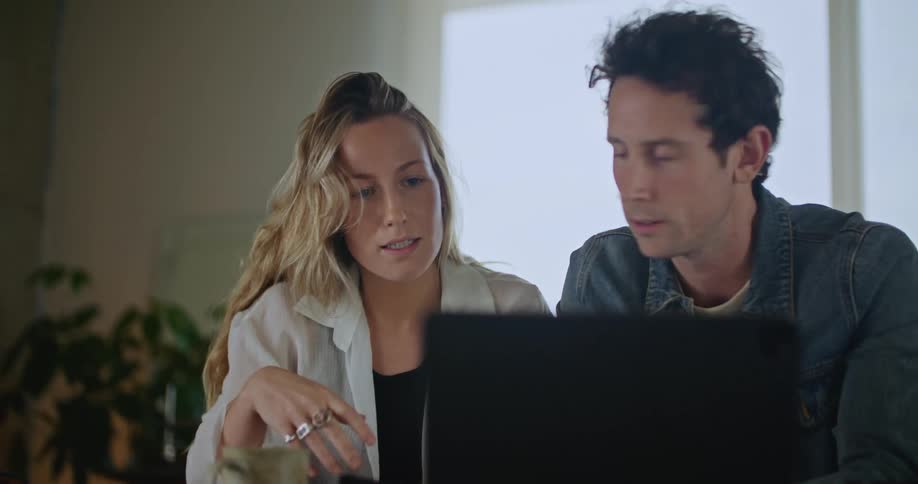 A man and a woman are sitting behind a laptop computer talking as she dials a number on her cell phone