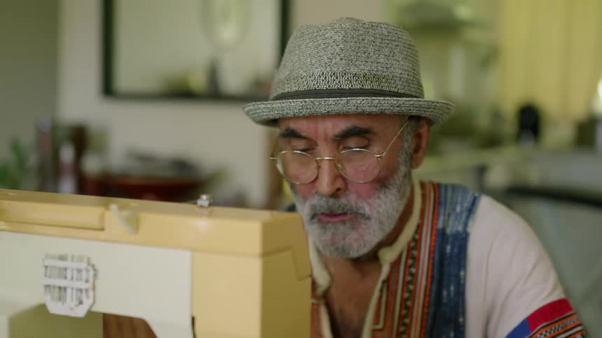 A man wearing a hat is sitting at a sewing machine holding fabric and guiding it under the needle
