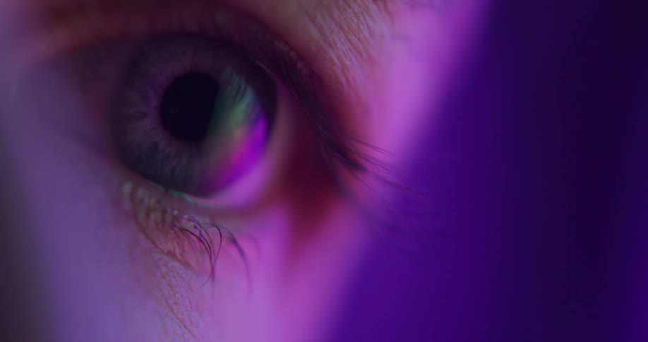 An extreme closeup of a persons eye as they look at something in a dark room full of colorful neon lights