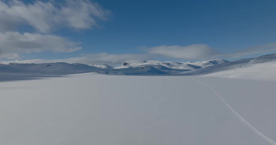 An aerial of a snow covered flat land with mountains in the far distance and a trail going through the snow