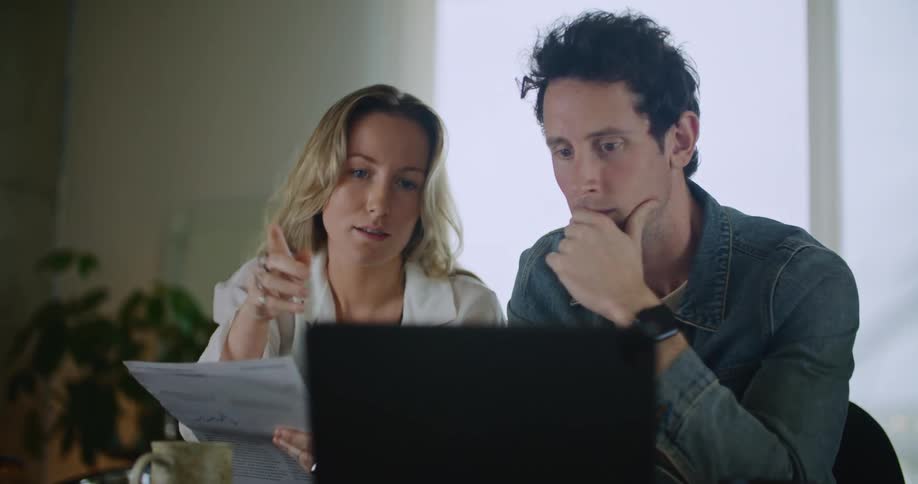 A man and woman talk while looking at a laptop at a table indoors during the day