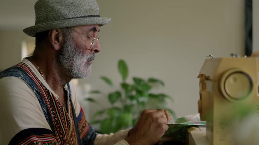 An elderly man is sewing clothes inside of his home