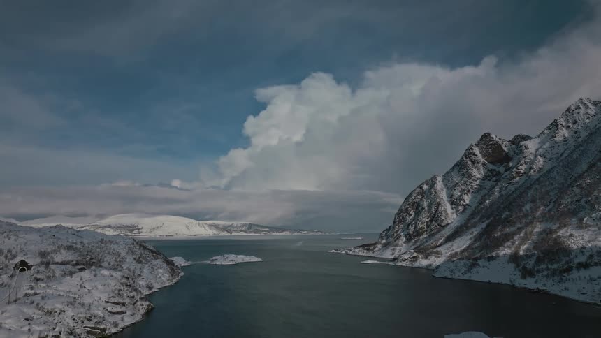 An aerial view of a river between two snow covered mountains with white fluffy clouds in the sky