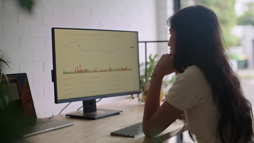 A young woman sitting at a desk uses a computer and monitor to analyze a line graph and bar graph