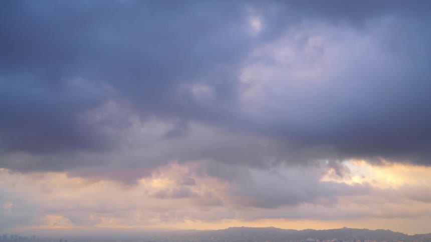 The sun shines on cumulus clouds as they move across a blue sky as the sun sets with mountains on the horizon