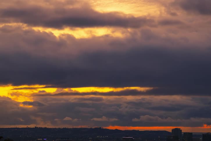 Clouds move over the city in a bright orange sky as the sun is setting and the colors are deeper with mountains on the horizon