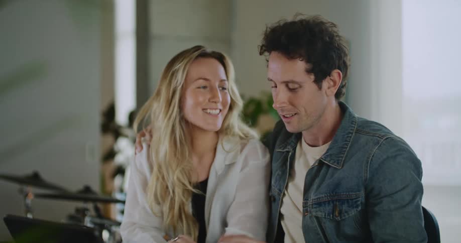 A man and woman sitting next to each other looking over documents as the man puts his arms around the woman as she puts her head down and laughs