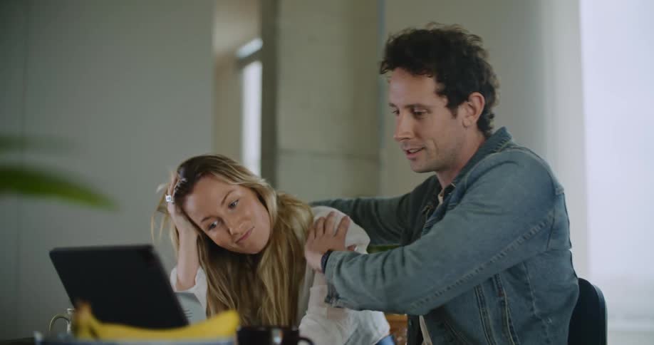 A man and woman sitting next to each other in front of papers and a laptop as they converse laugh smile and the man hugs the girl from the side