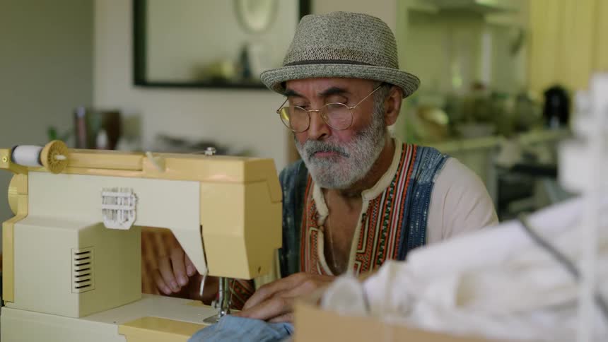 A man is sitting in his house and using a sewing machine