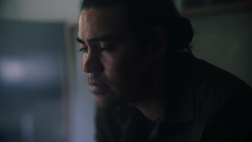 Light shines on a mans face as he stands in a smoke filled room looking ahead and blinking his eyes
