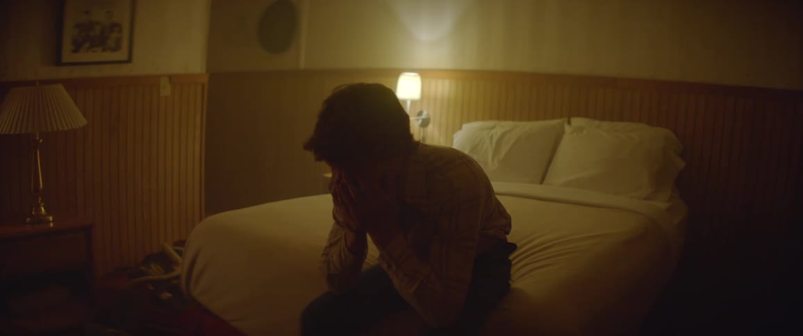 Man sits down on a bed with a single lamp lit in the room