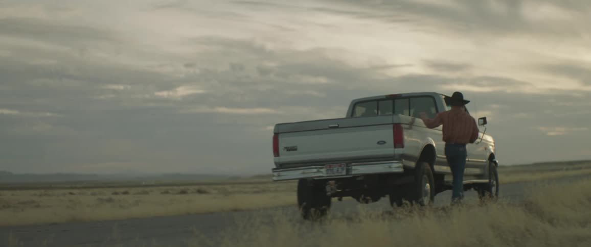 A pickup truck is slowing down to let a cowboy climb in on a cloudy day