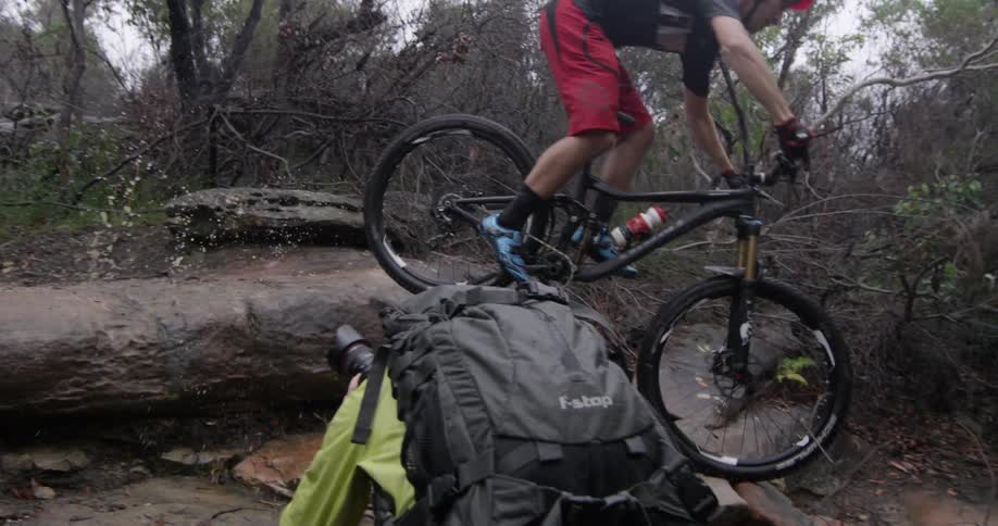 A man films a man riding a bike down a rocky path in the woods