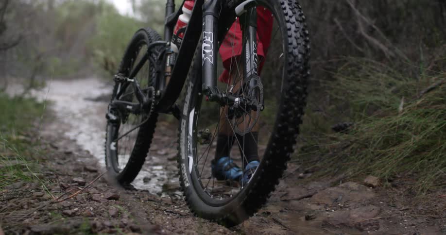 A man walks his bike over a bumpy muddy trail in the woods