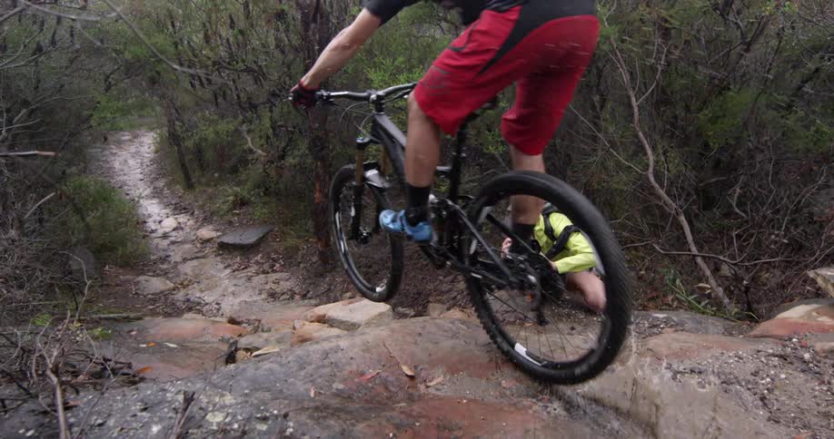 A man films a man riding a bike down a rocky path in the woods