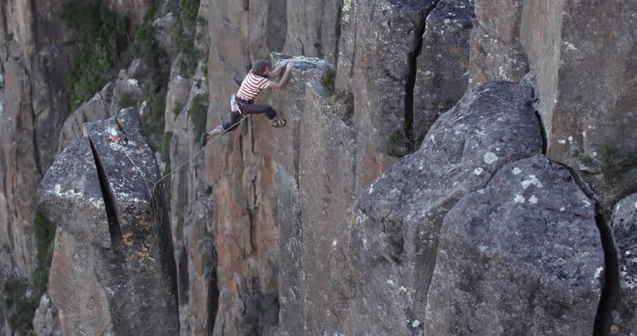 A rock climber leaps from a tall rock pedestal to the side of a cliff