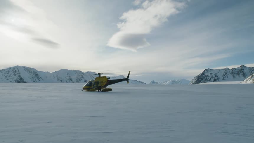 A man is standing near a helicopter sitting on the snow covered ground as the rotor spins and there are snow covered mountains in the distance