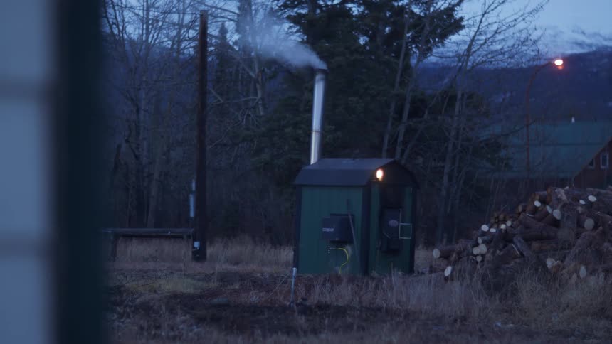 Small shack near a pile of wood with a chimney spewing white smoke in the countryside at evening