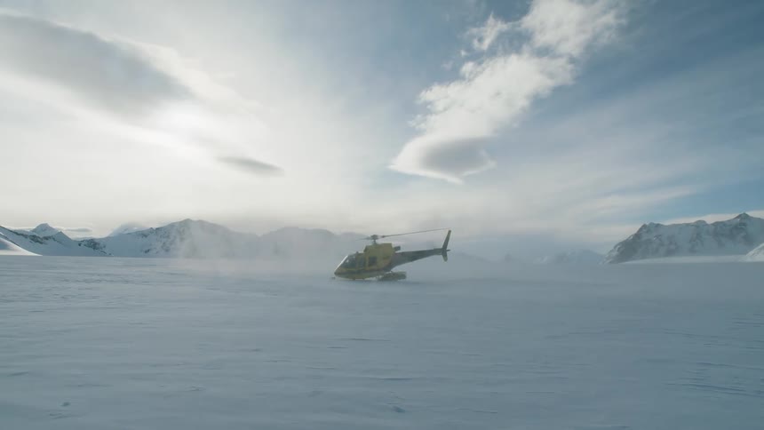 A helicopter is sitting on snow covered ground as the rotor spins and smoke flies in the air with mountains in the distance
