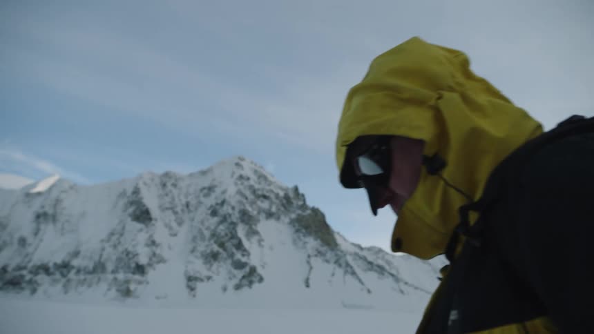 Mountaineer wearing a yellow jacket and goggles carries a backpack through the mountains