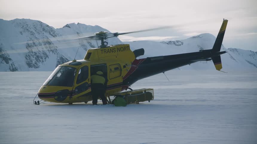 A helicopter is sitting on snow covered ground as the rotor spins and a man is taking supplies out and putting them on the ground