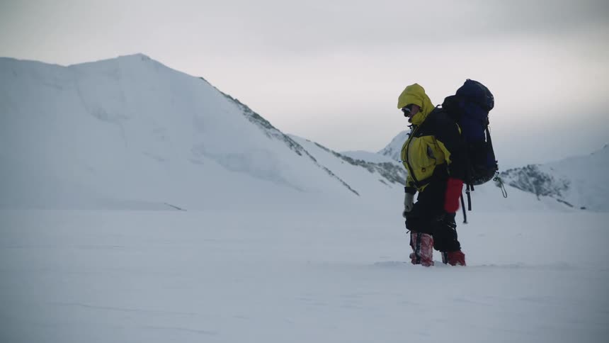 A backpacker struggles to hike through hard snow below a mountain range