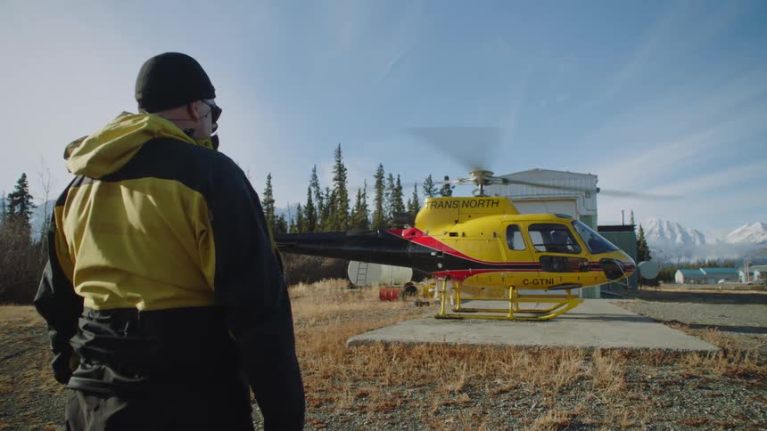 A helicopter spins its rotor on a helipad in front of mountains and a forest while a man watches
