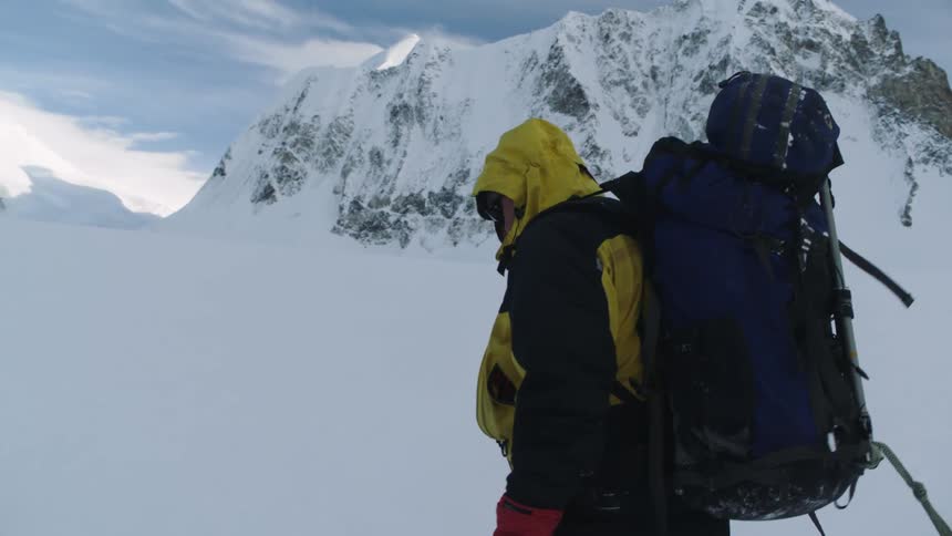 A person dressed for extreme weather slowly walks through deep snow in the mountains during the day