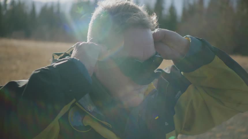 A man puts on mirrored goggles while sitting in a field near a forest