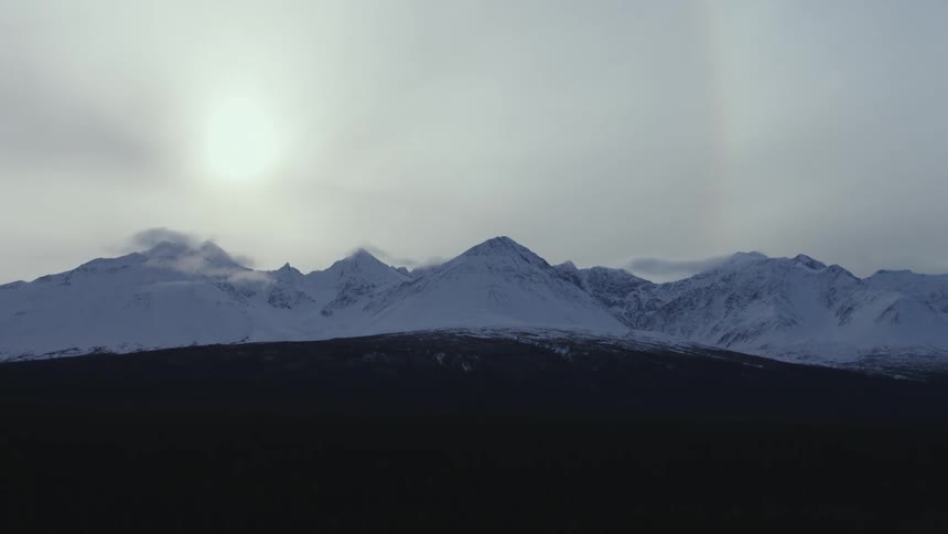 Panoramic view of the evening sun shining behind the clouds over the snow covered peaks of a mountain range