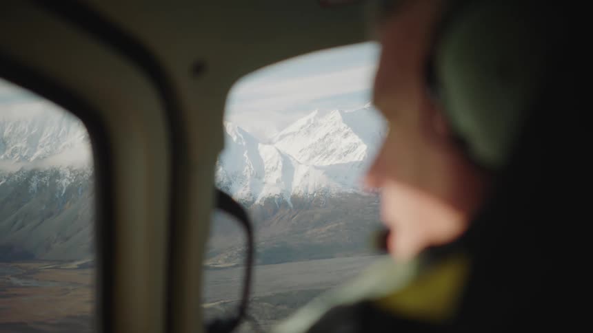 A man is wearing a headset is flying a helicopter near snow covered mountains as he looks out the window