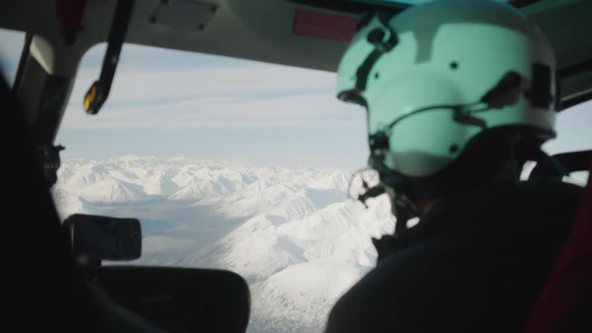 A pilot in the cockpit of an aircraft looks over and laughs as he flies over snow covered mountains