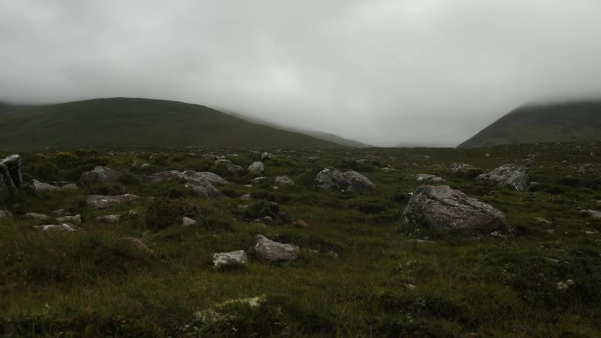 Large boulders laying on the grass in the hills near the mountains under low white clouds