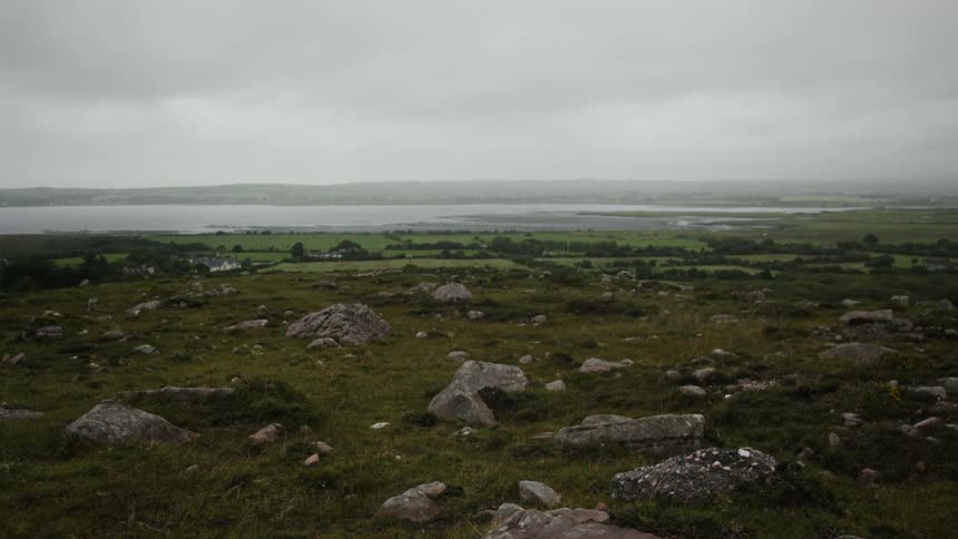 Wide open fields with rocks on the land and houses an ocean and mountains in the distance