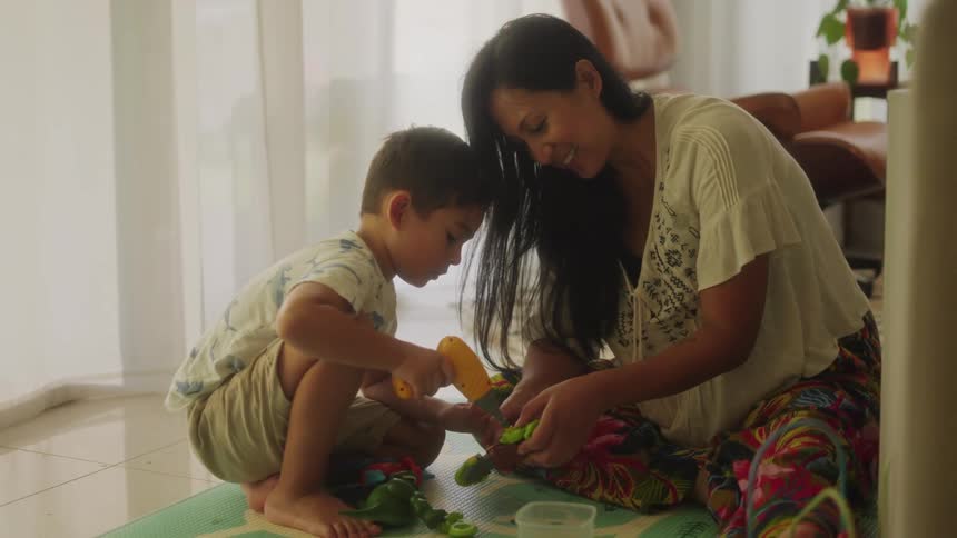 Mother with white shirt and colorful pants plays with her son on the floor in the living room at home